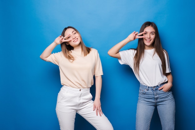 Picture of two playful girls standing together and showing peace gestures over blue wall