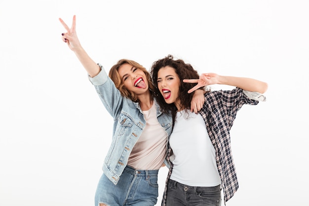 Picture of two playful girls standing together and showing peace gestures  over white wall