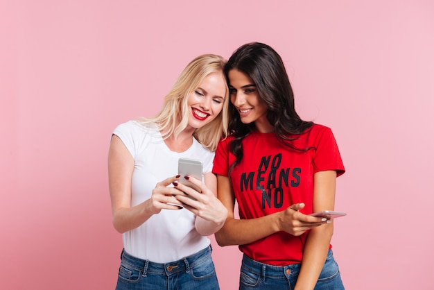 Free photo picture of two pretty happy women using her smartphones over pink