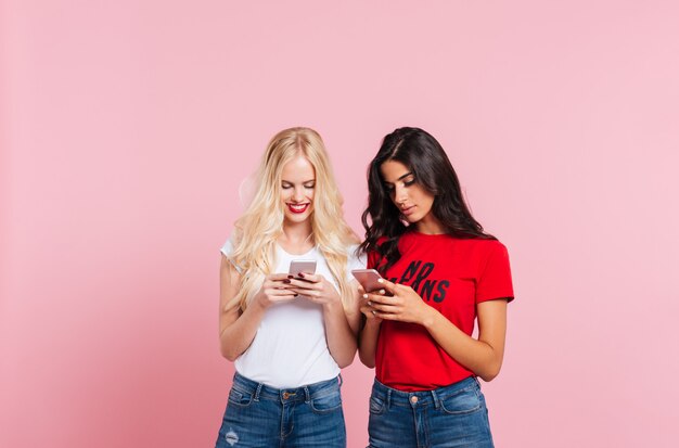 Изображение двух симпатичных улыбающихся женщин, использующих ее смартфоны на розовом