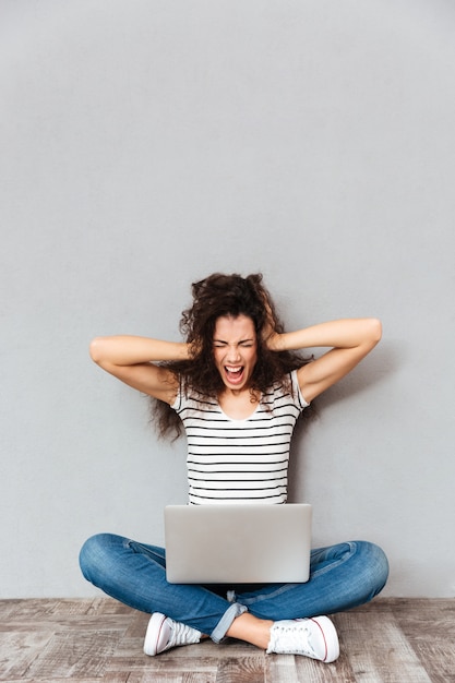 Picture of woman sitting with legs crossed on the floor screaming and covering ears being disappointed with her exam results over grey wall