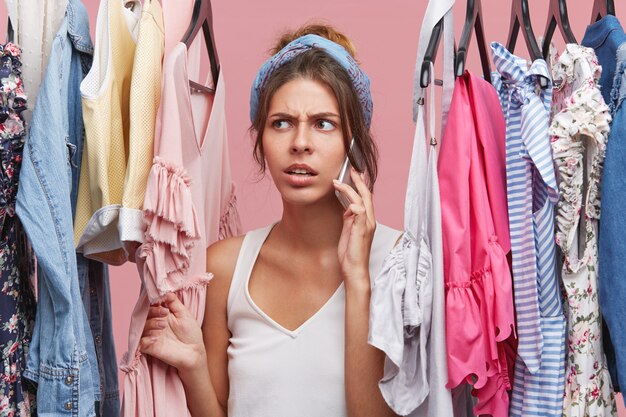 Picture of worried beautiful young Caucasian woman standing at wardrobe among summer clothes and talking on mobile phone to friend asking to help her while choosing perfect dress to wear on date