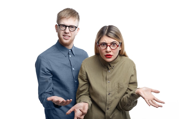 Picture of young bearded man and blonde woman both wearing glasses standing and expressing indignation, shrugging and making helpless gesture as they have no clue what is going on