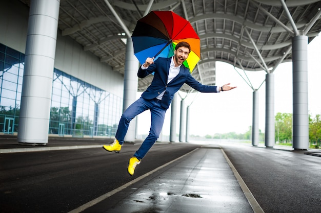 Picture of young businessman holding motley umbrella jumping and having fun at station