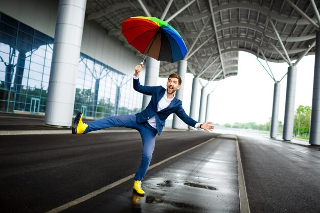 Изображение молодого бизнесмена, держащего пестрый зонтик, прыгающего и веселящегося на вокзале