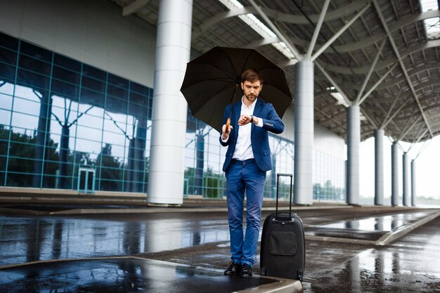 Картина молодой бизнесмен, холдинг чемодан и зонтик, глядя на часы, ожидание на дождливую станцию