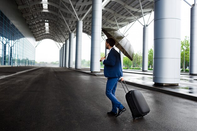 雨のターミナルでスーツケースと傘を保持している青年実業家の画像