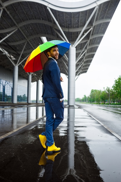 Picture of  young businessman in yellow shoes holding motley umbrella in rainy street