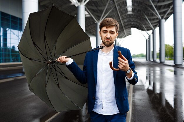 Picture of young confused businessman on street  holding broken umbrella