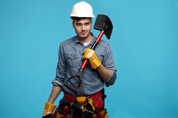 Picture of young digger wearing gloves, white helmet and tool belt carrying shovel