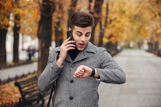 Изображение молодого парня, говорящего по smartmobile, глядя на часы, опаздывая