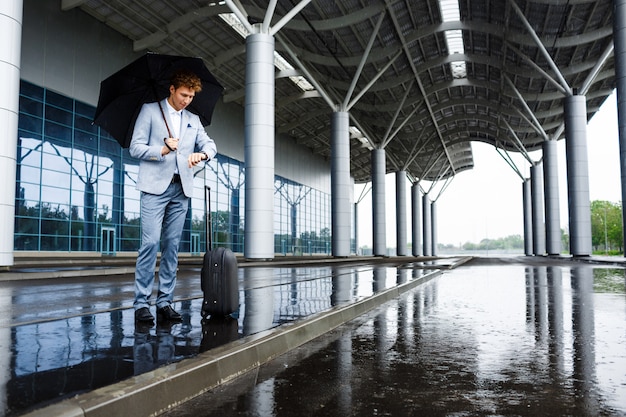 Picture of  young redhaired businessman holding black umbrella in rain and looking on watch at station