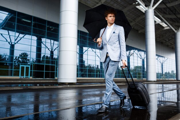 Picture of  young redhaired businessman holding black umbrella and suitcase walking in rain at airport