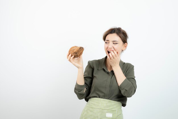 Picture of young woman in apron holding a coconut against white wall . High quality photo