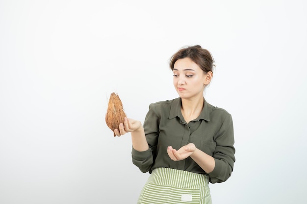 Picture of young woman in apron holding a coconut against white wall . High quality photo