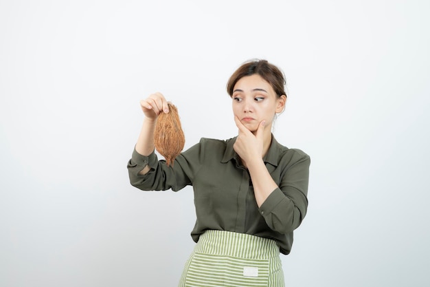 Picture of young woman in apron holding a coconut against white wall . High quality photo