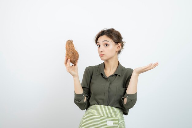 Picture of young woman in apron holding a coconut against white wall . High quality photo