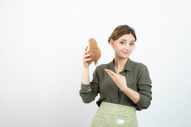 Picture of young woman in apron holding a coconut against white wall . High quality photo