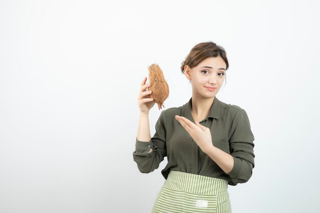 Picture of young woman in apron holding a coconut against white wall . High quality photo