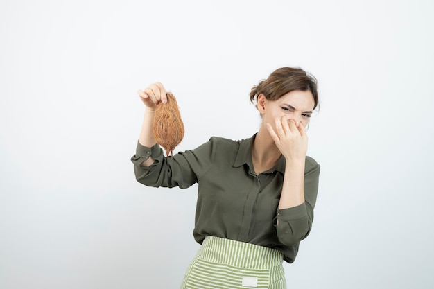 Immagine della giovane donna in grembiule che tiene una noce di cocco e che copre il naso. foto di alta qualità