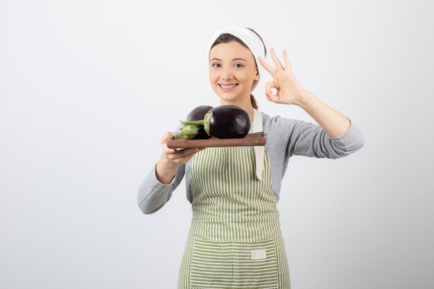 Picture of a young woman holding a wooden plate with eggplants and showing ok gesture