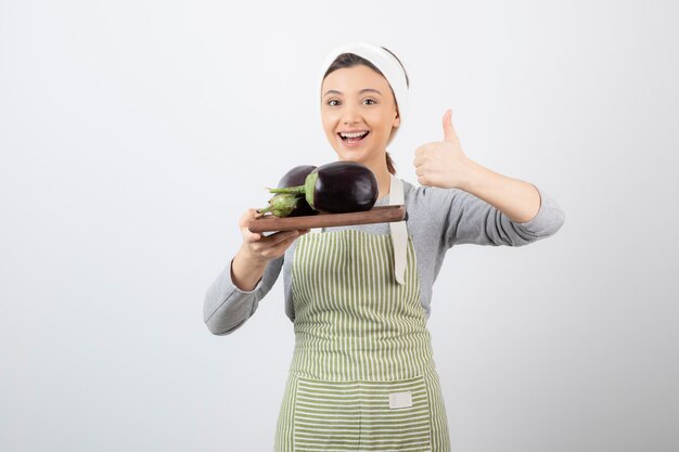 Picture of a young woman holding a wooden plate with eggplants and showing a thumb up