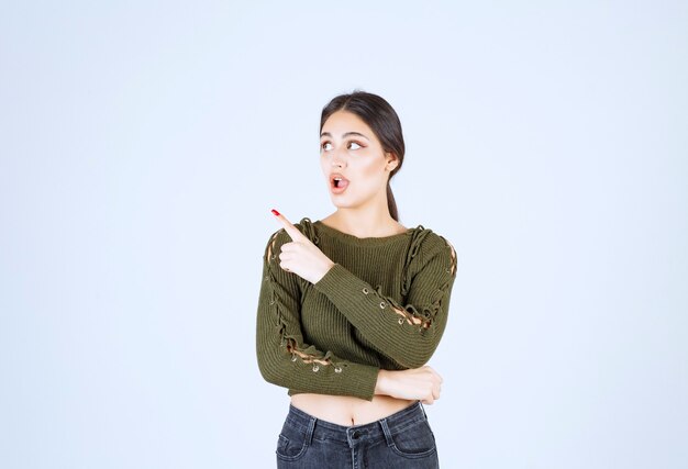 Picture of a young woman model standing and pointing aside with index finger .