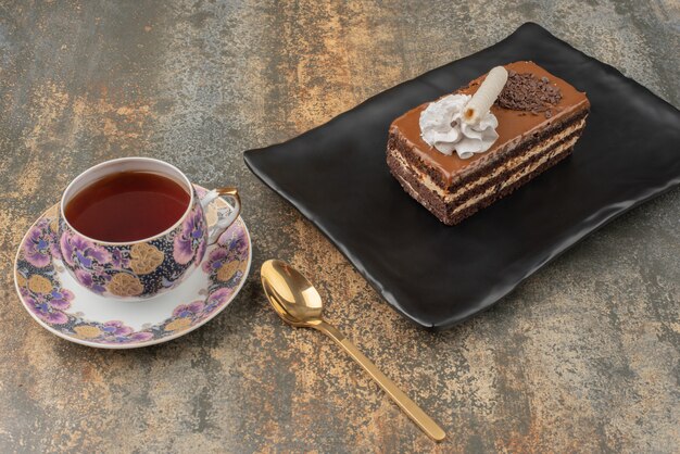 A piece of cake with hot tea and spoon on dark plate