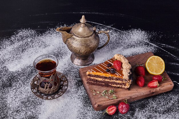 A piece of chocolate cake decorated with fruits on dark background with classic tea set . 