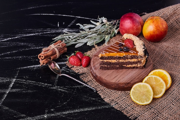 A piece of chocolate cake on round wooden plate with cinnamon and various fruits. 