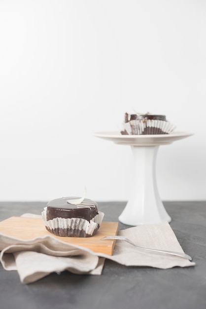 Free photo a piece of chocolate cake with wrapper on chopping board and cake stand on grey desk