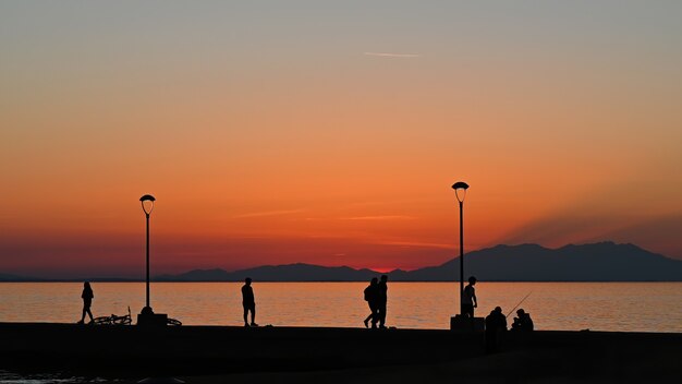 Pier with multiple fishing and walking people at sunset, parked bike, land  lampposts, Greece