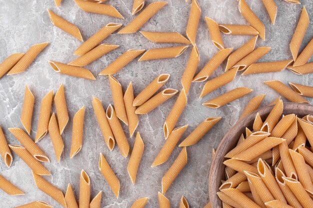 Pile of brown penne pasta placed in a wooden bowl on marble surface.