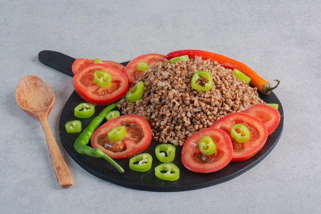 A pile of buckwheat topped with pepper and tomato on a wooden board next to a spoon on marble surface