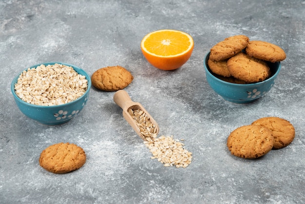 Foto gratuita mucchio di biscotti e farina d'avena in una ciotola e arancia tagliata a metà sulla superficie grigia.