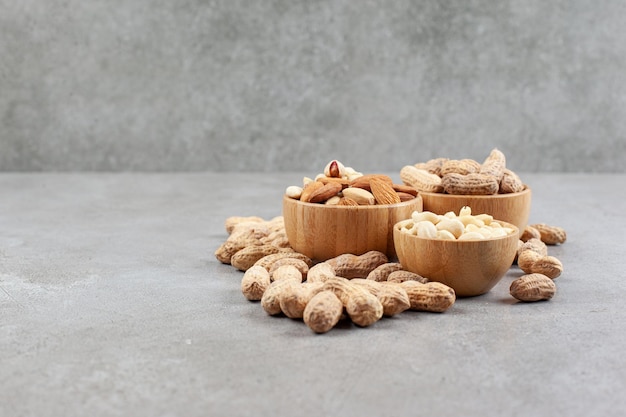 A pile of different nut types in bowls next to scattered peanuts on marble background. High quality photo