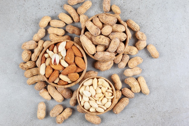 A pile of different nut types in bowls next to scattered peanuts on marble surface.