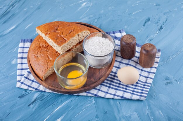 A pile of sliced bread on a board next to egg and flour on a tea towel , on the blue surface.