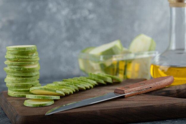 Pile of sliced zucchini on wooden board with knife and oil bottle.