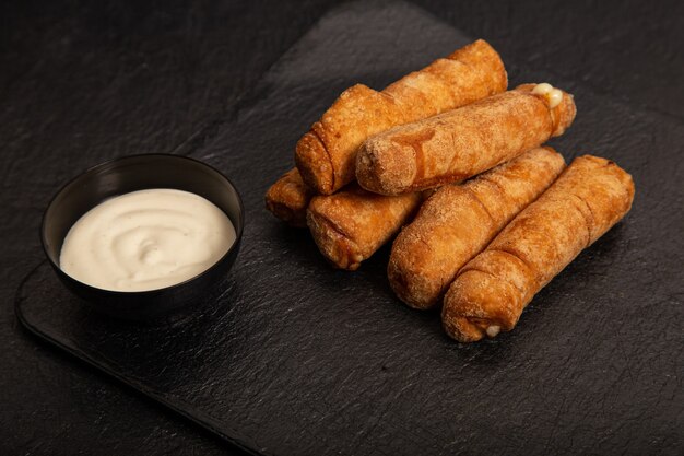 Pile of some fried rolls and a bowl of white sauce on the black background
