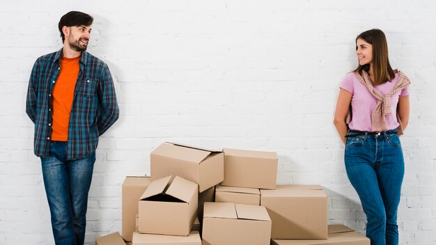 Piles of cardboard boxes between the stylish young couple standing against wall looking to camera