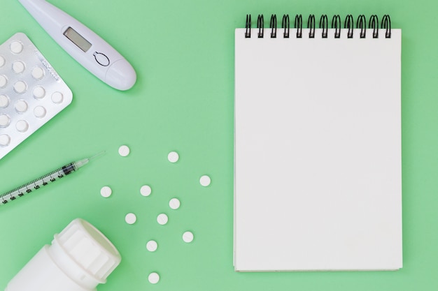 Pills; thermometer; syringe; plastic bottle near the spiral notepad on green background