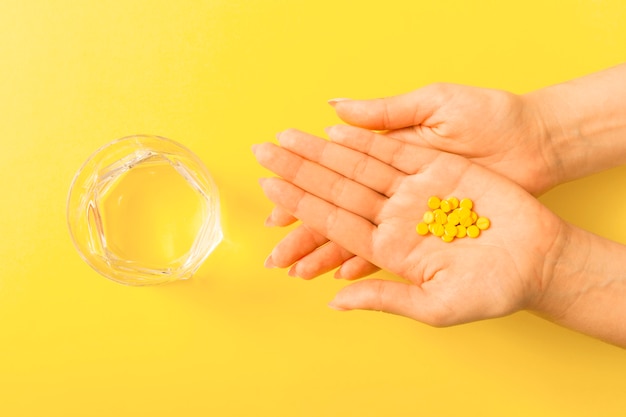 黄色の背景の上に水のガラスで女性の手の上の丸薬