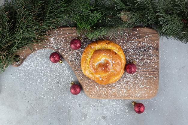 Pine branches, wooden board, christmas tree toys and a small bun on marble surface