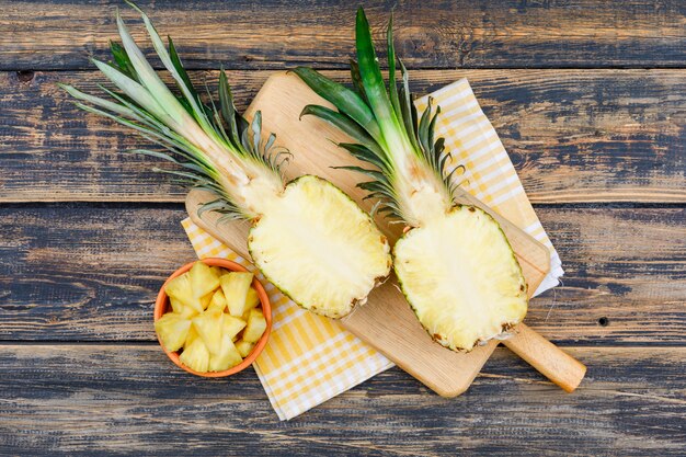 Pineapple halves and slices in a clay bowl and cutting board on old wood grunge surface and picnic cloth, flat lay.