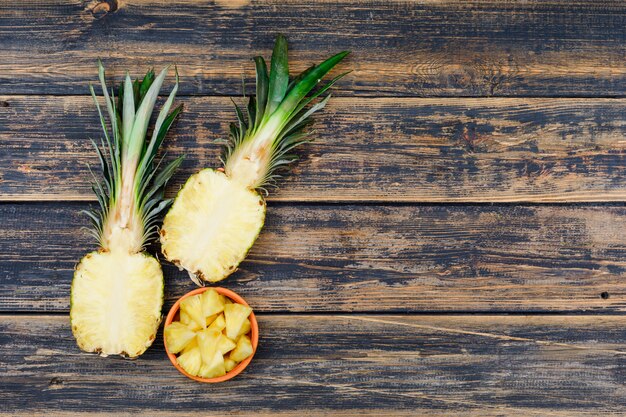 Pineapple halves and slices in a orange bowl on old wood grunge surface, top view.