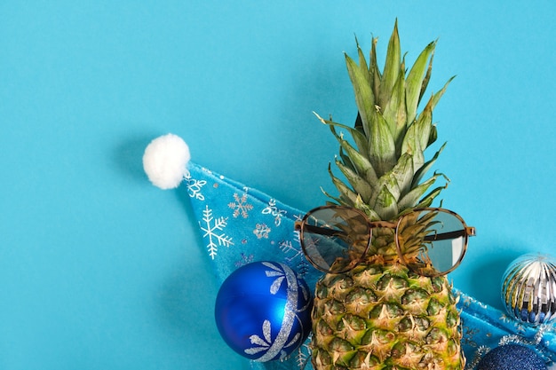 Pineapple in santa hat and sun glasses, christmas decor on blue background, winter vacation in tropics concept
