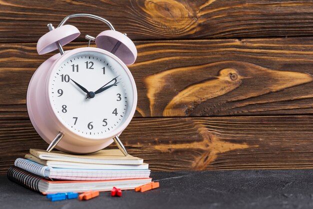 Pink alarm clock over the stacked of notebooks and clothes peg against wooden wallpaper