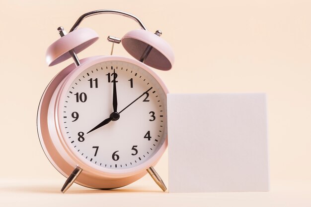 Pink alarm clock with blank white adhesive note on beige background