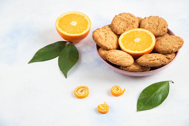 Pink bowl full with homemade cookies and half cut orange with leaves over white surface.